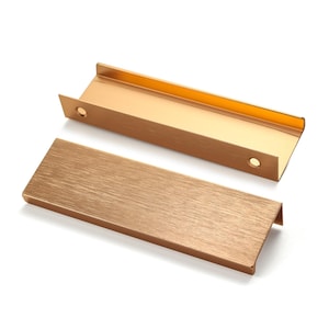 Könnte beinhalten: Zwei rechteckige Schrankgriffe mit gebürstetem Gold-Finish. Jeder Griff verfügt über eine flache, längliche Vorderseite mit texturierter Metallmaserung und einen rechtwinkligen Montageflansch mit zwei runden Schraublöchern.