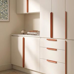 Puede incluir: Un conjunto de armarios de color blanco roto con largos tiradores de metal en tono cobre, dispuestos vertical y horizontalmente. La unidad de almacenamiento incluye puertas altas y tres cajones apilados con acabado mate liso.