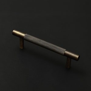 May include: A cylindrical cabinet handle with a dark bronze finish. The central section features a textured, diamond-patterned knurled grip, while the two ends and the two mounting posts have a smooth, brushed metallic surface finish.