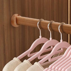 Puede incluir: Una barra de armario de madera con varias perchas de terciopelo rosa que sostienen suéteres color crema. Las perchas tienen ganchos plateados y están bien organizadas. El interior del armario es de madera.