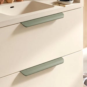 Op de afbeelding: Een crèmekleurig badkamermeubel met twee lades voorzien van lange, matte saliegroene horizontale handgrepen. Op het aanrecht staan drie kleine geometrische vetplantpotjes, een donker glazen parfumflesje, een decoratieve steen en een zeepbakje.