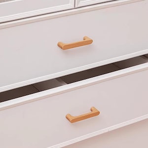 May include: Two light brown wooden drawer pulls with a smooth, curved design attached to a white dresser with framed drawer fronts. The top drawer is closed, while the bottom drawer is slightly open, revealing the interior space of the furniture.