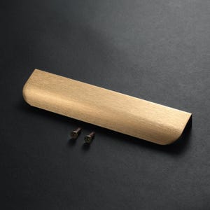 May include: A brushed brass cabinet pull with a curved edge and two small screws. The hardware has a rectangular shape with a textured surface and a warm, metallic finish. The pull is designed for easy installation.