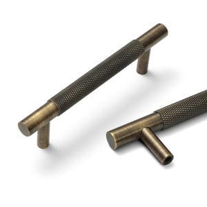 May include: Two cylindrical cabinet pulls with a dark antique brass finish. Each handle features a central section with a textured diamond knurled pattern and smooth, rounded ends. The pulls are mounted on two short, perpendicular cylindrical posts.