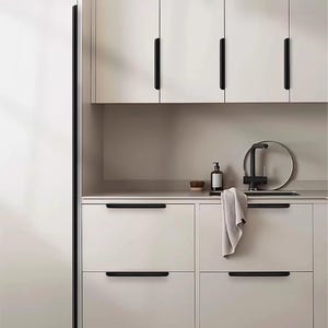 May include: A kitchen interior featuring off-white cabinetry with matte black linear handles. The setup includes a black tap, a soap dispenser, a small wooden bowl, and a light-coloured cloth draped over a drawer handle, set against a neutral wall.