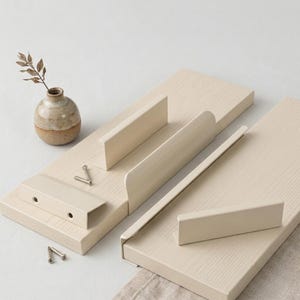 Puede incluir: Dos estantes flotantes de madera de color beige claro con una textura de veta de madera visible. Cada estante cuenta con una barandilla protectora de metal beige a juego en el borde frontal. Dos tornillos metálicos pequeños están sobre el estante.