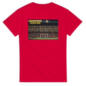 Può includere: T-shirt rosso con una grafica raffigurante uno stadio di calcio e la scritta "There will only ever be one CLOCK END". L'immagine mostra una partita di calcio in corso con giocatori in campo e tifosi sugli spalti. La maglietta è in materiale morbido.