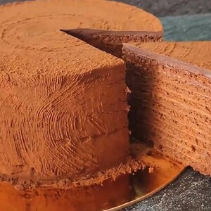 Puede incluir: Un pastel de chocolate redondo de varias capas cubierto con un fino polvo de cacao. El exterior presenta un acabado texturizado y arremolinado. Se ha retirado una porción triangular, revelando las finas capas apiladas de pastel y glaseado.