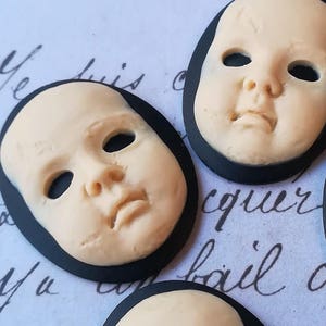 May include: Five oval-shaped doll face charms with black borders. The doll faces are made of a cream-colored material and have painted eyes, noses, and mouths.