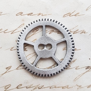 May include: A silver-colored gear with a circular design and multiple spokes. The gear has small teeth around the outer edge and is set against a backdrop of aged paper with handwritten script.