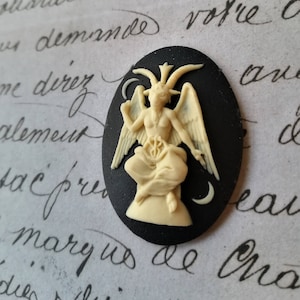 May include: A black cameo featuring a white, detailed sculpture of a winged figure with horns and a pentagram. The figure is seated on a crescent moon.