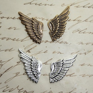 Op de afbeelding: Vier metalen vleugelbedels, twee in brons en twee in zilver, met gedetailleerde veermotieven. De bedels liggen op een crèmekleurige papieren achtergrond met handschrift, wat een vintage uitstraling geeft.