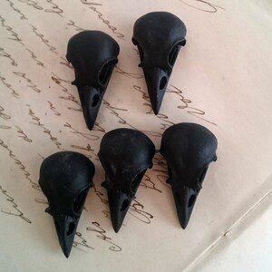 May include: Six black crow skull charms, each with a detailed design, are arranged on a piece of paper with handwritten text.