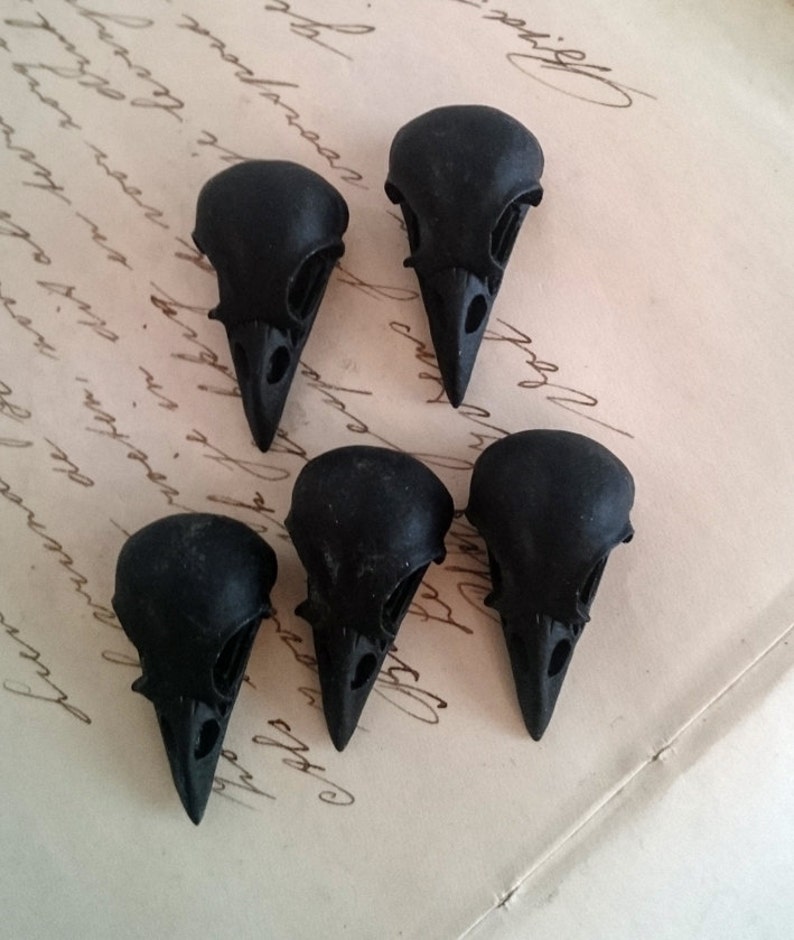 May include: Six black crow skull charms, each with a detailed design, are arranged on a piece of paper with handwritten text.