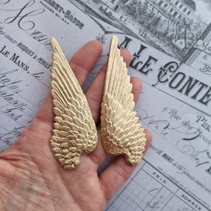 May include: Two gold-coloured metal angel wings with detailed feather designs. The wings are held in a hand against a background of a vintage document.