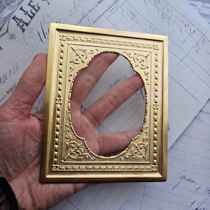Puede incluir: Un marco de fotos de metal dorado con un diseño ornamentado. El marco tiene una forma rectangular con un recorte ovalado decorativo en el centro.