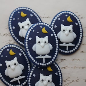 Pode incluir: Cinco botões ovais azuis e brancos com um design de coruja branca. Cada botão apresenta uma coruja branca com olhos amarelos, uma lua crescente amarela e estrelas brancas sobre um fundo azul escuro.