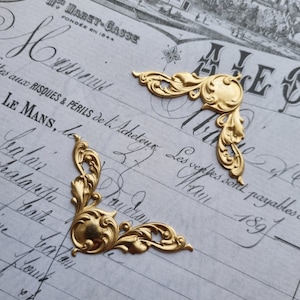 May include: Two ornate, gold-colored metal corner embellishments. Each corner piece features a circular center with swirling leaf and floral designs. The pieces rest on a vintage paper background with faded script and text.