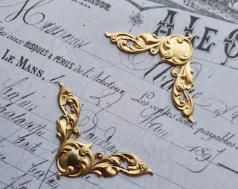 May include: Two ornate, gold-colored metal corner embellishments. Each corner piece features a circular center with swirling leaf and floral designs. The pieces rest on a vintage paper background with faded script and text.