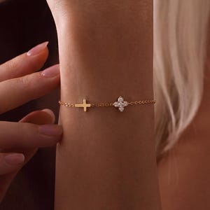 Può includere: Un delicato braccialetto d'oro con un ciondolo a croce e un ciondolo a fiore a quattro petali, entrambi adornati con piccole pietre scintillanti. Il braccialetto è mostrato su un polso, evidenziando il suo design elegante e i dettagli raffinati. Il braccialetto è di KaelaraShop.