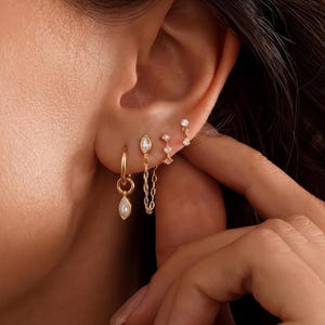 Könnte beinhalten: Ein Set goldfarbener Ohrringe, bestehend aus einem kleinen Creolen-Ohrring mit marquiseförmigem Anhänger, einem Kettenohrring mit Marquise-Stein, einem vertikalen Stab-Ohrring mit kleinen Steinen und einem einzelnen Ohrstecker mit klarem Stein.