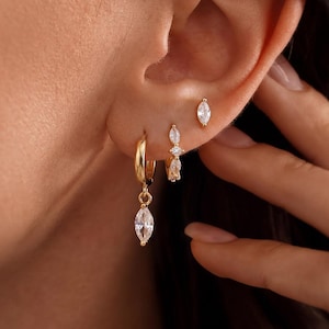Puede incluir: Un conjunto de tres pendientes dorados con piedras transparentes de corte marquesa. El primero es un aro pequeño con un dije colgante, el segundo es una barra vertical con tres piedras pequeñas y el tercero es un pendiente de botón individual.