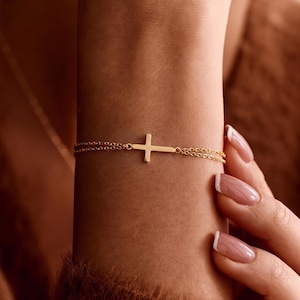 May include: A delicate gold-toned bracelet featuring a horizontal cross charm centred on a double-strand fine chain. The minimalist design showcases a polished metallic finish, with the cross positioned horizontally across the wrist area.