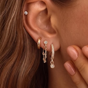 Könnte beinhalten: Ein Set goldfarbener Ohrringe, bestehend aus einem kleinen Stecker am oberen Ohr, einem schlichten Creolen-Ohrring, einem Kettenglied-Ohrring mit kleinem runden Stein und einem mit Zirkonia besetzten Creolen-Ohrring mit einem hängenden runden Stein.