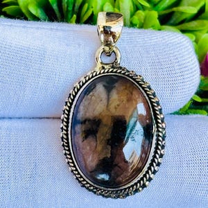 May include: Oval pendant with a polished, multi-toned gemstone in shades of brown and tan. The stone is set in a gold-colored metal frame with a rope-like border and a matching bail.