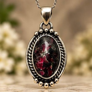 May include: An oval pendant featuring a dark, mottled stone with deep red and black inclusions, set in a polished silver frame with a twisted rope border and small spherical accents, suspended from a silver chain link necklace.