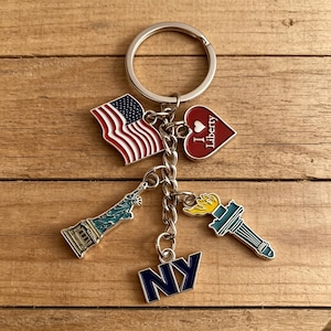 May include: A silver keychain with multiple charms. Charms include an American flag, a red heart with "I Liberty" text, a Statue of Liberty, a torch, and "NY" in blue. The charms are attached to a silver ring.