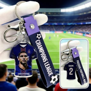 May include: A keychain with a soccer jersey design, featuring the text "ACHRAF HAKIMI" and "CHAMPIONS LEAGUE". The keychain has a blue and purple strap with a soccer ball design.