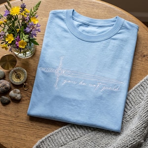 May include: A light blue t-shirt with a white sword design and the text "you do not yield." The shirt is folded on a wooden surface, next to a vase of flowers, a compass, and a gray knitted blanket.