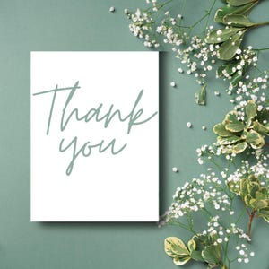 Puede incluir: Una tarjeta blanca con las palabras "Thank you" en una suave escritura verde. La tarjeta se encuentra sobre un fondo verde apagado, adornada con delicadas flores blancas y follaje verde, creando una est&eacute;tica natural y elegante.