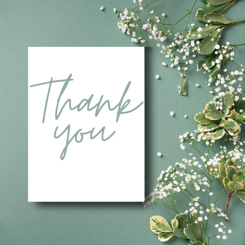 Puede incluir: Una tarjeta blanca con las palabras "Thank you" en una suave escritura verde. La tarjeta se encuentra sobre un fondo verde apagado, adornada con delicadas flores blancas y follaje verde, creando una est&eacute;tica natural y elegante.