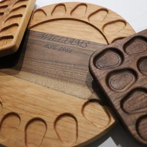 May include: A circular light wood serving board with a dark wood center strip engraved with the text WILLIAMS EST. 1966, featuring carved indentations along the perimeter, accompanied by a rectangular light wood tray and a square dark wood tray.