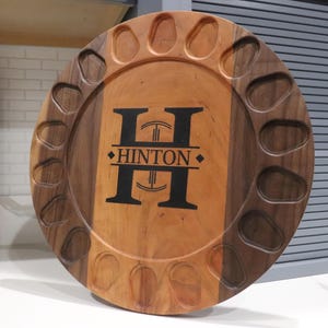 May include: A round wooden serving board featuring a central light wood panel flanked by dark wood sides. The board has a circular perimeter with twelve carved indentations. The center displays a large black letter H with the text HINTON.