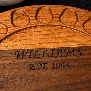 May include: A circular wooden serving board featuring a two-tone design with a lighter wood top section containing carved indentations and a darker wood bottom section engraved with the text WILLIAMS EST. 1966 in a serif font.