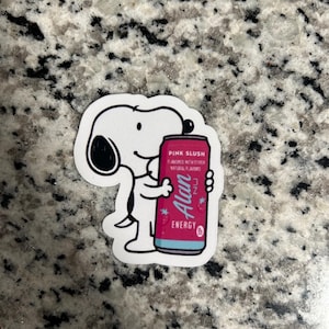May include: A white sticker featuring a cartoon dog holding a pink energy drink can. The can is labeled "Pink Slush" and "Alan Energy" with other text. The sticker has a white border and is on a speckled surface.