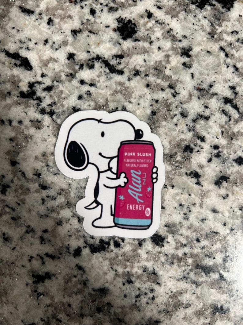 May include: A white sticker featuring a cartoon dog holding a pink energy drink can. The can is labeled "Pink Slush" and "Alan Energy" with other text. The sticker has a white border and is on a speckled surface.