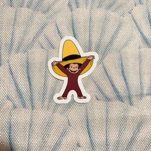 May include: A die-cut sticker featuring a brown monkey character wearing a large yellow hat with a black band. The monkey has its arms raised, holding the brim of the hat, and is smiling. The sticker has a white border and a smooth, matte finish.