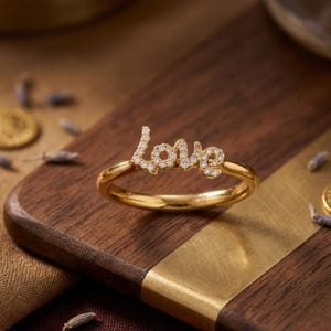 May include: A gold ring spelling out "Love" in a cursive font, embellished with small, sparkling stones. The ring is displayed on a wooden surface with a gold accent, with sprigs of lavender and a gold coin scattered around.