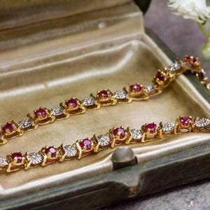 Op de afbeelding: Een gouden armband met afwisselend rode edelstenen en heldere, ronde stenen. De armband wordt getoond in een open, groene vintage sieradendoos met een gouden satijnen binnenkant. Witte bloemen zijn op de achtergrond.