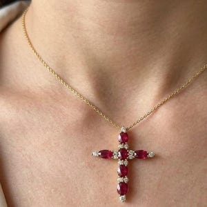 Pode incluir: Um colar dourado com um pendente de cruz. A cruz é adornada com pedras preciosas ovais vermelho-escuras e pequenas pedras preciosas redondas e claras. O colar é exibido em um fundo cor de pele.