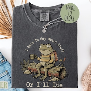 May include: Dark gray t-shirt with a frog graphic. The frog is sitting on a log, holding a coffee cup, and wearing a backpack. Text on the shirt reads "I Have To Say Weird Stuff Or I'll Die". A silver necklace is on the shirt.