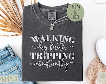Może przedstawiać: Ciemnoszary t-shirt z krótkim rękawem i okrągłym dekoltem, ozdobiony białym nadrukiem WALKING by faith TRIPPING constantly. Koszulka posiada metkę marki Comfort Colors przy wewnętrznej stronie kołnierza i jest prezentowana ze srebrnym naszyjnikiem.