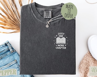 Puede incluir: Una camiseta de manga corta gris carbón con un gráfico blanco en el pecho. El gráfico muestra un libro abierto rodeado de pequeñas estrellas y el texto "JUST ONE MORE CHAPTER". La camiseta tiene cuello redondo y una etiqueta de marca.