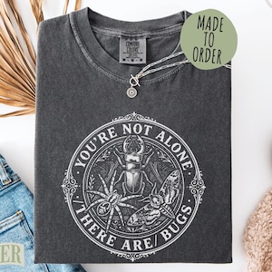 May include: Dark gray t-shirt with a circular graphic featuring a beetle, spider, and moth, with the text "YOU'RE NOT ALONE / THERE ARE / BUGS." A silver necklace and a "MADE TO ORDER" sticker are visible. The shirt is folded on a white surface.