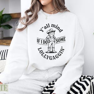 White sweatshirt with a frog wearing a cowboy hat and overalls, text reads "Y'all mind if I do some lollygaggin'".