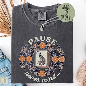 May include: A charcoal gray t-shirt featuring a graphic print of a mahjong tile with a peacock illustration, surrounded by a circular wreath of orange flowers and dark leaves. The text reads PAUSE above and never mind... below the design.